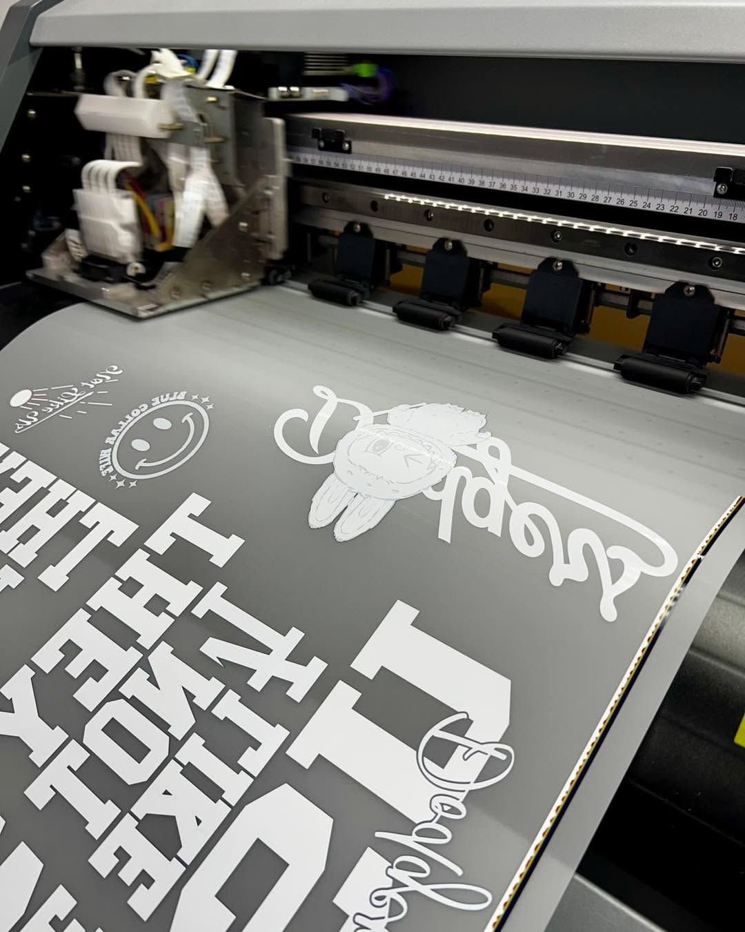Large format printer laying down a white vinyl decal design on gray material